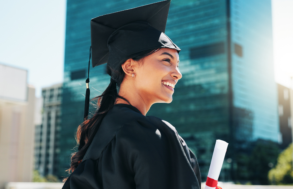 A smiling graduate in a cap and gown holding a diploma, with city buildings in the background.